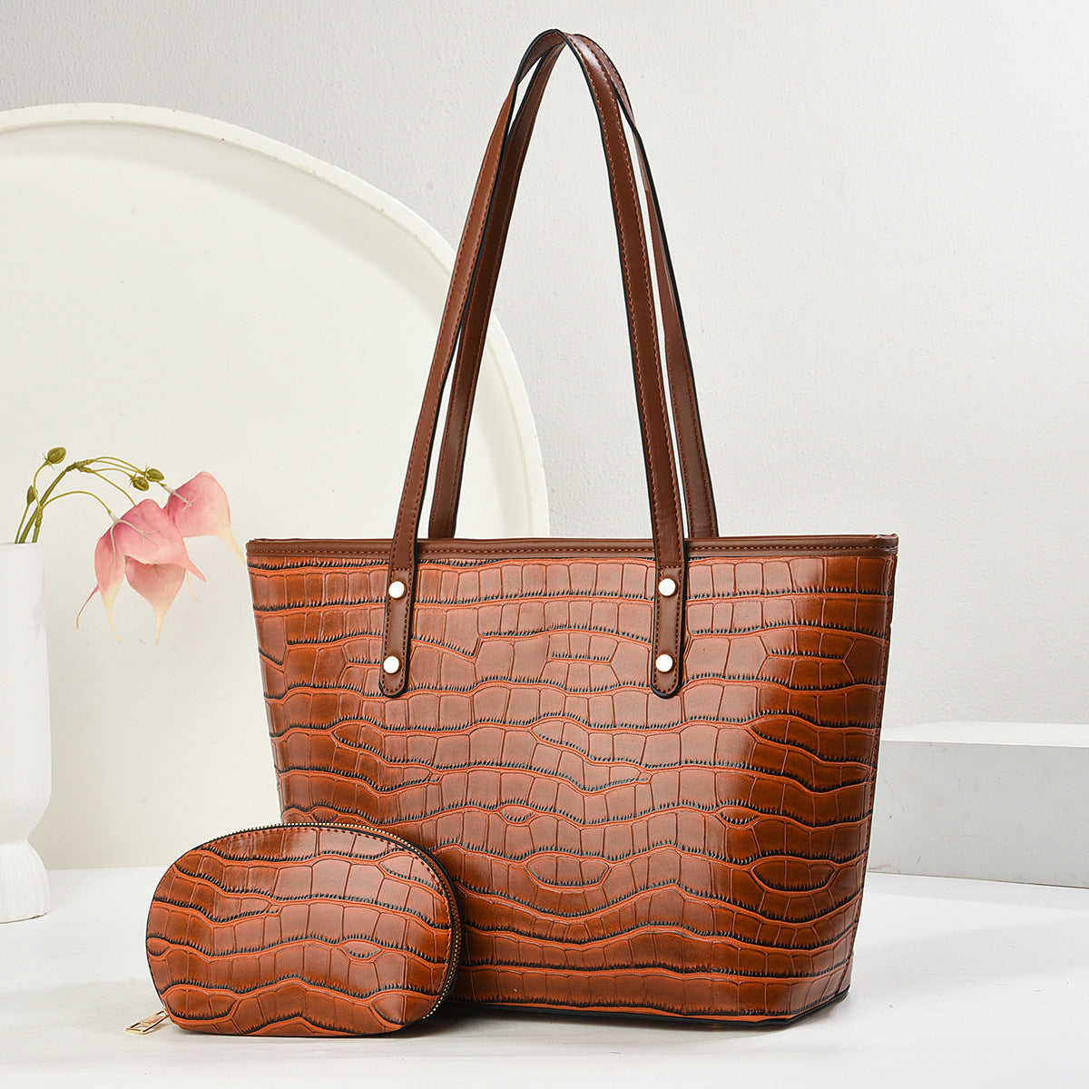 A brown crocodile-embossed leather tote bag with matching shoulder straps and a matching zippered coin pouch, displayed on a white surface against a white background with a decorative vase.